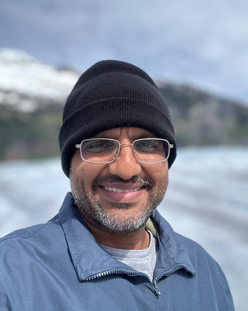 Portrait of Amar P. Patel wearing a beanie and glasses, smiling, with a glacier and mountains in the background
